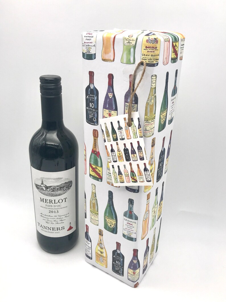 May include: A white wine bottle gift bag with a colorful pattern of various wine bottles. The bag has a brown twine bow and a small gift tag with a similar pattern. A bottle of red wine is next to the bag. The bottle label reads "Merlot Pays d'Oc 2013 Tanners Established 1945".