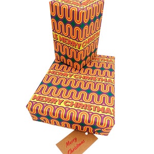 May include: Two wrapped gifts with a repeating wavy pattern in yellow, purple, green, and orange. The words "MERRY CHRISTMAS" are printed across the wrapping paper. A small brown tag with the words "Merry Christmas" hangs from the bottom gift.