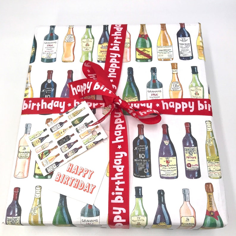 May include: Red and white gift wrap with a pattern of wine bottles. The gift wrap is tied with a red ribbon with the text "happy birthday" repeated on it. A small white gift tag with the text "Happy Birthday" is attached to the ribbon.