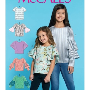 May include: A sewing pattern for girls' tops with ruffled sleeves. The pattern includes five different styles: a striped top, a floral print top, a solid purple top, a floral print top with a butterfly design, and a green polka dot top. The pattern is labeled "McCall's M7799".