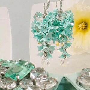 Può includere: Un paio di orecchini pendenti con una cascata di fiori in pasta polimerica blu chiaro e dettagli in cristallo.