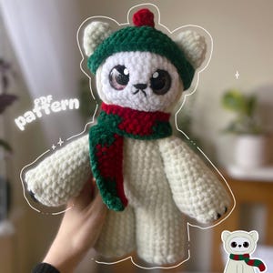 May include: A handmade white crochet bear wearing a green hat with a red pom-pom and a red and green scarf. The bear has large, expressive eyes and a small black nose. The text "PDF pattern" is written on the left side of the image.