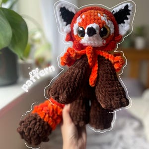 May include: A handmade crochet red panda plushie with a vibrant orange and brown color scheme. The plushie features large, expressive eyes, a fluffy tail, and the text "pdf pattern". The plushie is held in a person's hand.