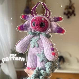 May include: A handmade crochet doll in shades of purple, pink, and green. The doll has large, glittery eyes, a round head, and a whimsical design. The text "PDF pattern" is visible on the left side of the image.