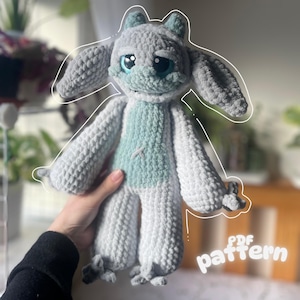 May include: A handmade crochet plush toy in shades of gray and light blue. The toy has large, expressive blue eyes, long ears, and a light blue belly. The text "PDF pattern" is visible in the lower right corner. The toy is held in a person's hand.