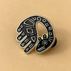 May include: Gold-toned enamel pin featuring a black and white stylized design of a creature with a curled tail and a hand-like shape.