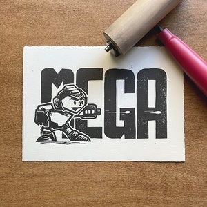 May include: Black and white illustration of a robot character with the word "MEGA" printed in bold letters. The robot is holding a weapon and is facing the right side of the image.