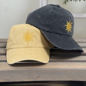 May include: Two baseball caps, one in a muted mustard yellow and one in a faded charcoal black, each featuring a small, embroidered yellow sun design on the front panel. The caps have a structured crown, a curved brim, and a classic six-panel design.
