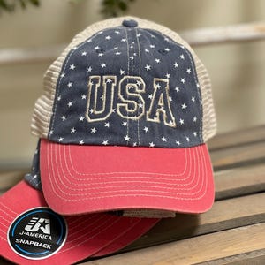 Könnte beinhalten: Eine Baseballkappe mit dunkelblauem Vorderteil, weißen Sternen und dem in Gold gestickten Text USA. Die Kappe hat einen roten Schirm und beigefarbene Netz-Seitenteile. Ein runder Aufkleber auf dem Schirm zeigt die Aufschrift J-AMERICA SNAPBACK mit Logo.