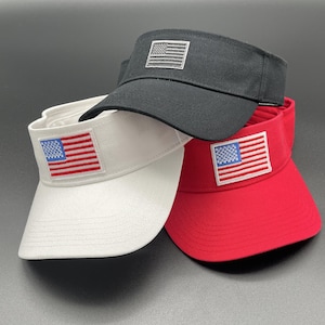 May include: Three sun visors in black, white, and red. Each visor features a rectangular patch on the front panel. The black visor has a grey-toned American flag patch, while the white and red visors display full-colour American flag patches.