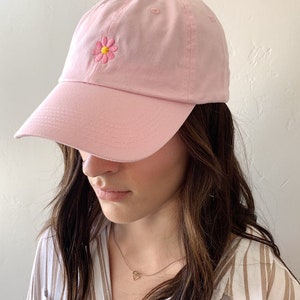 May include: A light pink baseball cap with a curved brim. The cap features a small, embroidered daisy with pink petals and a yellow center on the front. The cap is made of a soft fabric and has a rounded crown.
