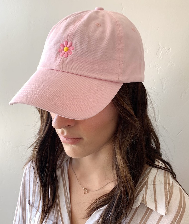 May include: A light pink baseball cap with a curved brim. The cap features a small, embroidered daisy with pink petals and a yellow center on the front. The cap is made of a soft fabric and has a rounded crown.