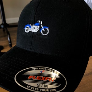 Könnte beinhalten: Eine schwarze Baseballkappe mit einer gestickten blau-weißen Motorradgrafik auf der Vorderseite. Der Schirm zeigt einen silbernen Aufkleber mit dem Text THE ORIGINAL FLEXFIT S/M IT FITS YOUR LIFE in schwarzer, oranger und blauer Schrift.