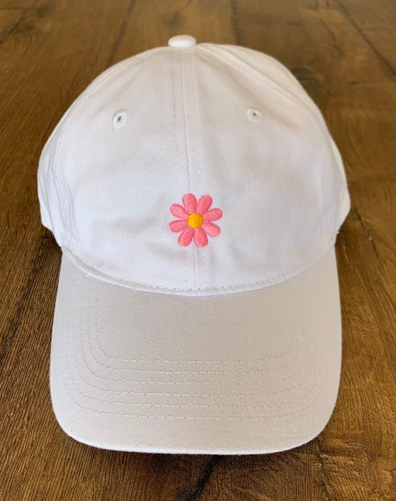 May include: White baseball cap with a curved brim and a pink and yellow daisy embroidered on the front. The cap has a button on top and eyelets for ventilation. The brim is a lighter shade of white.