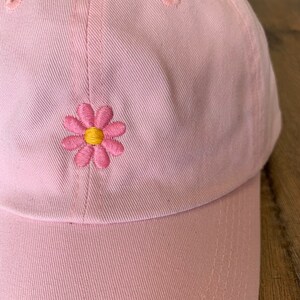 May include: A light pink baseball cap with a stitched daisy flower design. The daisy has pink petals and a yellow center. The cap is made of a soft fabric and has a curved brim.
