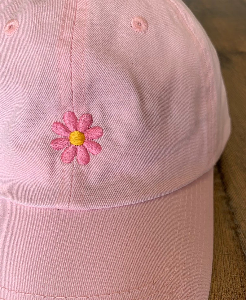 May include: A light pink baseball cap with a stitched daisy flower design. The daisy has pink petals and a yellow center. The cap is made of a soft fabric and has a curved brim.
