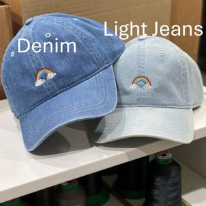 May include: Two baseball caps, one in medium blue denim and one in light blue denim, each featuring an embroidered rainbow and white cloud design. The text Denim is positioned above the darker cap, and Light Jeans is above the lighter cap.