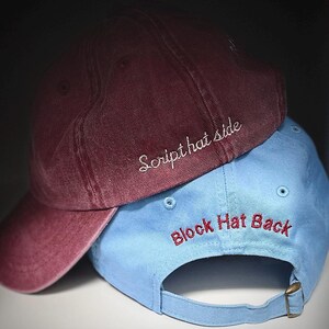 May include: Two baseball caps, one burgundy and one light blue, stacked on top of each other. The burgundy cap has the text "Script hat side" embroidered in white. The light blue cap has the text "Block Hat Back" embroidered in red.