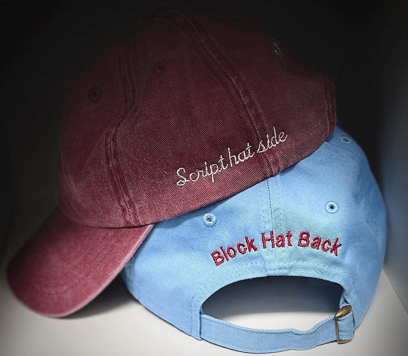May include: Two baseball caps, one burgundy and one light blue, stacked on top of each other. The burgundy cap has the text "Script hat side" embroidered in white. The light blue cap has the text "Block Hat Back" embroidered in red.