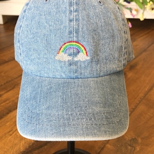 Puede incluir: Una gorra de béisbol de mezclilla azul con un arcoíris y nubes bordadas en la parte delantera.