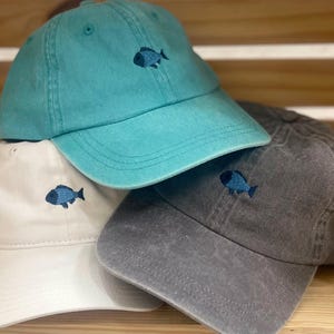 May include: Three baseball caps in teal, off-white, and charcoal gray. Each cap features a small, embroidered dark blue fish icon centered on the front panel. The hats have a structured crown, a curved brim, and visible stitching details.