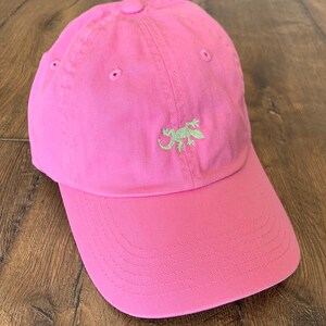 Può includere: Un cappellino da baseball rosa acceso con un piccolo geco ricamato in verde chiaro al centro del pannello frontale. Il cappellino presenta una corona strutturata, una visiera curva con cuciture a vista e occhielli rosa coordinati.