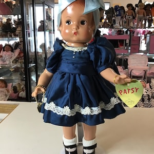May include: A blue and white dress with lace trim, worn by a porcelain doll with a blue bow in her hair. The doll is holding a small purse and has a tag that reads "Patsy, An Effanbee Doll".