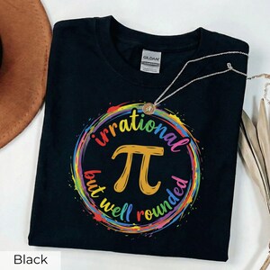 May include: Black t-shirt with a colorful graphic design featuring the mathematical symbol Pi, surrounded by the words "irrational but well rounded." A silver necklace with a small pendant rests on the shirt.