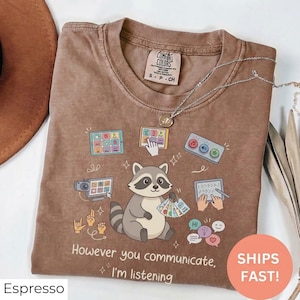 Könnte beinhalten: Espressofarbenes T-Shirt mit einer Waschbär-Grafik, die Kommunikationskarten hält. Das Shirt zeigt den Text "However you communicate, I'm listening" und verschiedene Kommunikationssymbole. Marke Comfort Colors.