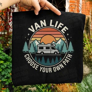Puede incluir: Bolso tote negro con un diseño gráfico circular que presenta una furgoneta camper, montañas y árboles. El texto "VAN LIFE" se arquea sobre la imagen, con "CHOOSE YOUR OWN PATH" debajo. El diseño tiene un esquema de colores retro.