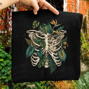 Könnte beinhalten: Ein schwarzes T-Shirt mit einem Grafikdruck eines menschlichen Brustkorbs und Herzens, durchwoben von grünen Monstera-Blättern, Farnen und orangefarbenen Paradiesvogelblumen. Das Motiv ist mittig auf dem Stoff platziert.