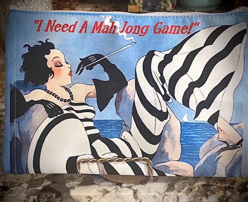 May include: A blue and white striped zippered pouch with a black and white illustration of a woman in a striped dress smoking a cigarette. The text "I Need A Mah Jong Game!" is printed on the pouch.