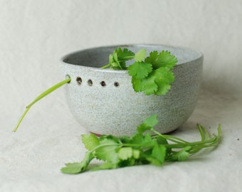 May include: A speckled, light gray ceramic bowl with a row of small holes near the rim. Fresh green cilantro sprigs are placed inside and next to the bowl, creating a natural, rustic aesthetic. The bowl has a slightly irregular shape.