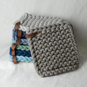 May include: A stack of handmade, square, knitted pot holders in various shades of blue, green, and grey. Each pot holder has a thick, braided edge and a brown leather tag with the text "BP Pottery".