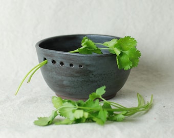 May include: A dark gray ceramic bowl with a series of small holes near the rim. Fresh green cilantro sprigs are arranged inside and around the bowl, creating a natural, rustic aesthetic. The bowl has a slightly rough texture and a simple, modern design.