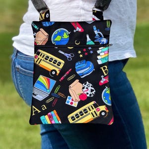 Könnte beinhalten: Eine schwarze Umhängetasche mit einem bunten Muster zum Thema Schule. Der Aufdruck zeigt gelbe Schulbusse, Globen, Laptops mit Regenbögen, Mikroskope, Scheren, Büroklammern, Bücher sowie die Buchstaben A und B auf einem dunklen Hintergrund.