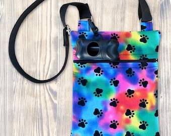 Puede incluir: Un bolso bandolera rectangular con un vibrante patrón tie-dye en tonos azul, rosa, amarillo y verde con un motivo de huellas negras. El bolso incluye una correa negra ajustable y un dispensador de bolsas de plástico negro con forma de hueso.