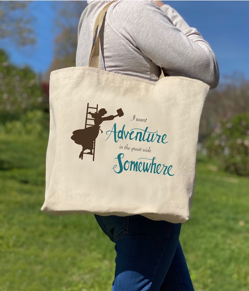 Op de afbeelding: Een beige canvas draagtas met een bruin silhouet van een persoon die een ladder beklimt en een mok vasthoudt. De tekst "I want Adventure in the great wide Somewhere" is gedrukt in teal. De tas heeft beige bandjes.