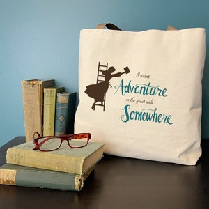 Op de afbeelding: Een cr&egrave;mekleurige draagtas met bruine handvatten, met een silhouet van een vrouw die een ladder beklimt. De turquoise tekst luidt "I want Adventure in the great wide Somewhere". Oude boeken en een rood montuur bril liggen ervoor.