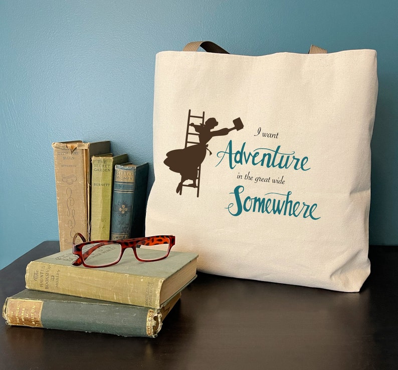 Op de afbeelding: Een cr&egrave;mekleurige draagtas met bruine handvatten, met een silhouet van een vrouw die een ladder beklimt. De turquoise tekst luidt "I want Adventure in the great wide Somewhere". Oude boeken en een rood montuur bril liggen ervoor.