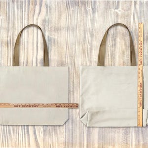 Op de afbeelding: Twee beige canvas draagtassen met bruine handvatten worden getoond tegen een houten achtergrond. Een houten liniaal is op elke tas geplaatst om de afmetingen te meten. De tassen zijn rechthoekig met een eenvoudig ontwerp.