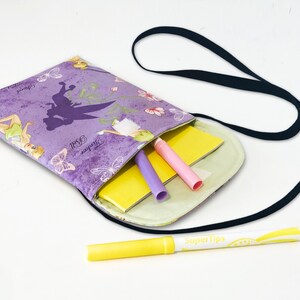 May include: Purple fabric crossbody bag with a Tinkerbell print and a black strap. The bag is open and has a yellow notebook, a pink highlighter, and a purple highlighter inside.