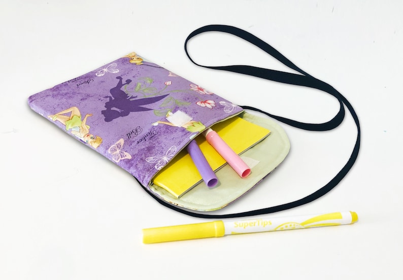 May include: Purple fabric crossbody bag with a Tinkerbell print and a black strap. The bag is open and has a yellow notebook, a pink highlighter, and a purple highlighter inside.