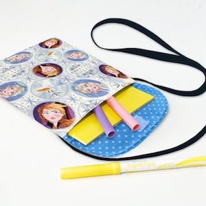 May include: A blue and white fabric bag with a black strap and a Frozen theme. The bag is filled with school supplies, including a yellow notebook, purple and pink pens, and a yellow marker.