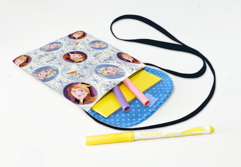 May include: A blue and white fabric bag with a black strap and a Frozen theme. The bag is filled with school supplies, including a yellow notebook, purple and pink pens, and a yellow marker.