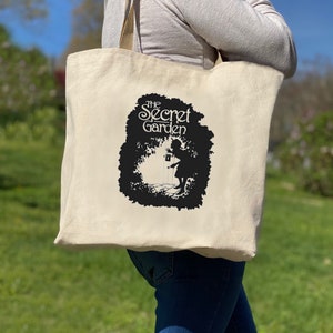 Peut inclure: Un sac fourre-tout beige avec une silhouette noire d'une fille tenant une lanterne devant un arbre. Le texte "The Secret Garden" est imprimé en noir au-dessus de la silhouette.