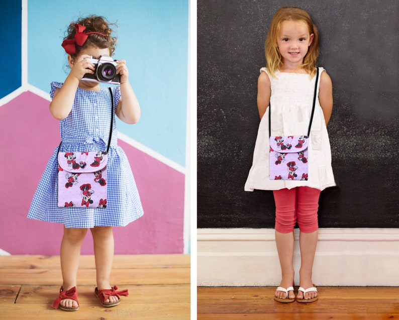 May include: Two children wearing colorful dresses and carrying small purple purses with a cartoon character print. The purses have adjustable straps and are worn crossbody.