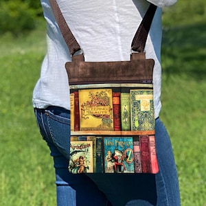 Peut inclure: Un sac bandoulière rectangulaire orné d'un imprimé de dos de livres anciens sur des étagères, incluant les titres Sleeping Beauty et The Lost Princess of Oz. Le sac possède une partie supérieure en tissu marron foncé, une fermeture éclair et une bandoulière réglable marron.