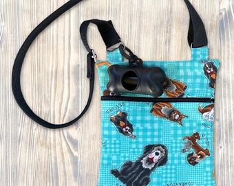 Puede incluir: Un bolso cruzado turquesa con una correa negra y un dispensador de bolsas para excrementos de perro negro. La bolsa presenta un patrón repetido de varias razas de perros y las palabras "Kisses", "Hugs", "Companion" y "Rescued". Una cremallera negra es visible.