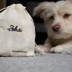 May include: A white canvas drawstring bag with the word "Zohsi" embroidered in black. The bag has a brown tag attached to it and the text "Kohsipets.com" printed on the bottom. A white dog with brown eyes is looking at the bag.